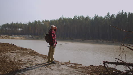 aged-fisher-is-viewing-reservoir-in-forest-standing-on-high-coast-at-sunny-autumn-day-angling-and-spin-fishing