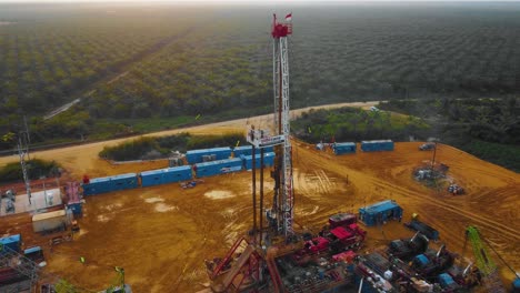 cinematic drone footage of onshore drilling rig equipment structure for oil exploration and exploitation in the middle of jungle surrounded by palm oil trees during sunset and high oil price