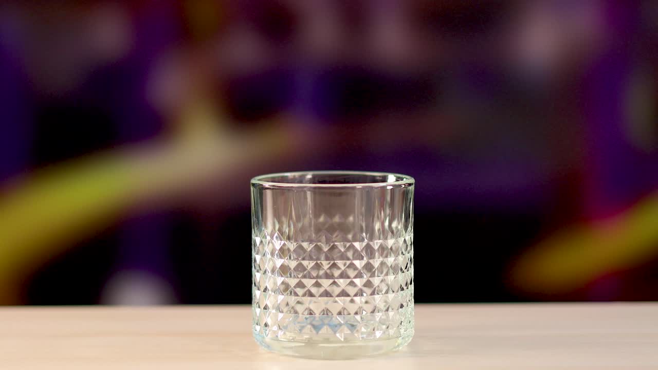 An ice cube is dropped into a textured glass on a wooden surface, set against a colorful blurred background