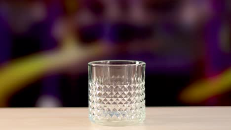 an ice cube is dropped into a textured glass on a wooden surface, set against a colorful blurred background