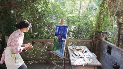 Artista-Birracial-Tomando-Fotos-De-Su-Pintura-Usando-Un-Teléfono-Inteligente-En-Un-Jardín-Soleado,-En-Cámara-Lenta