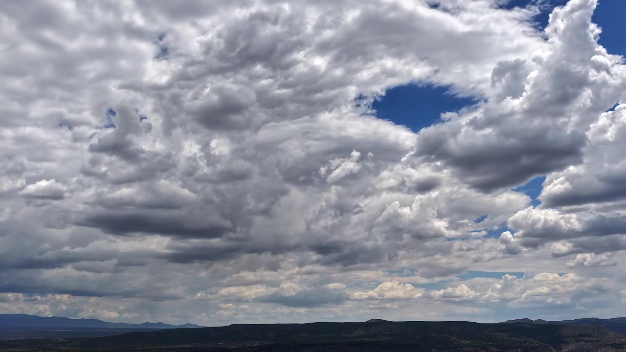 4K drone-timelapse, der fanger et smukt, vidtstrakt hav af hurtigt skiftende skyer over et mørkt, højt ørkenlandskab i sydvest. Pletter af klar blå himmel bryder igennem det tætte, rullende skylandskab.