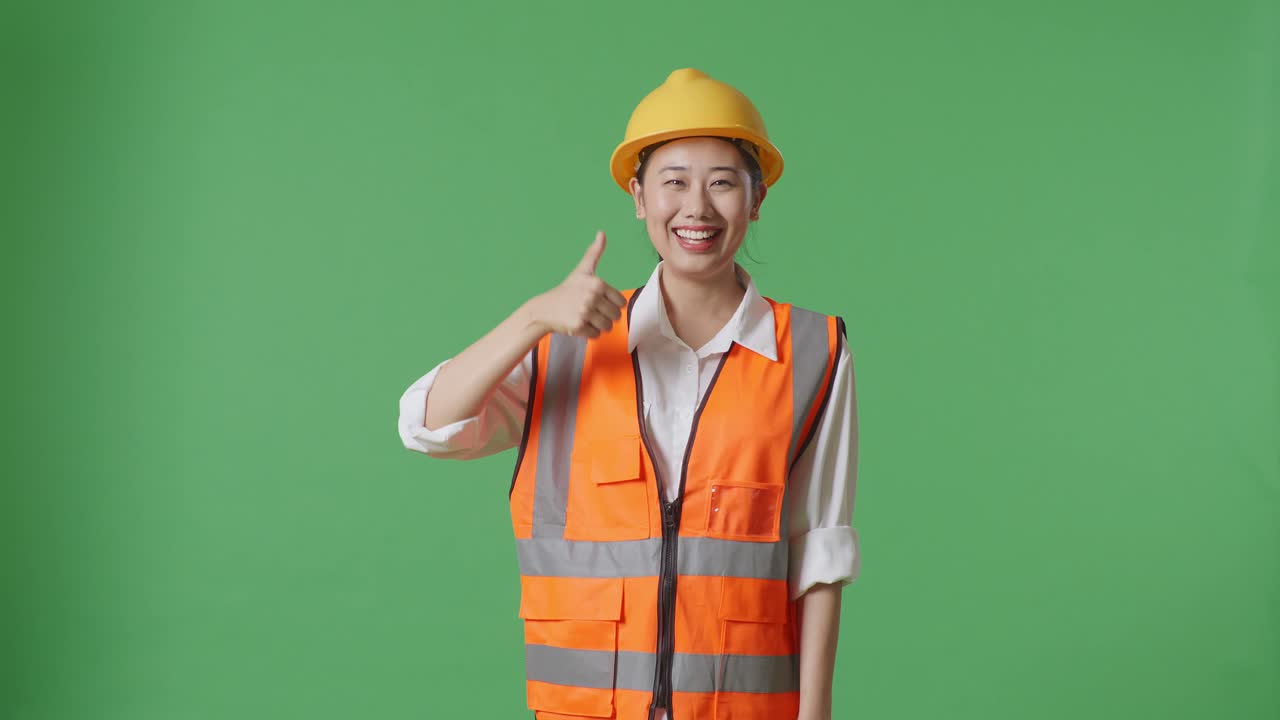 Asian Female Engineer With Safety Helmet Smiling And Showing Thumbs Up Gesture To The Camera While Standing In The Green Screen Background Studio