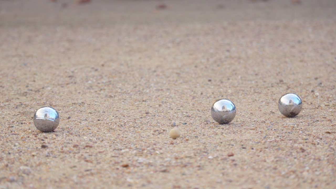 Slow-motion close-up of pétanque balls. One ball hits another, knocking it away from the cochonnet. Dust rises, capturing the precision of the strike in daylight.