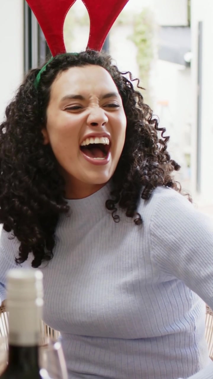 Vertical video: Laughing woman in blue top and red antler reacting to pal joke at table with wine