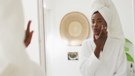 Video-of-happy-african-american-woman-in-robe-looking-at-mirror-and-moisturizing-face