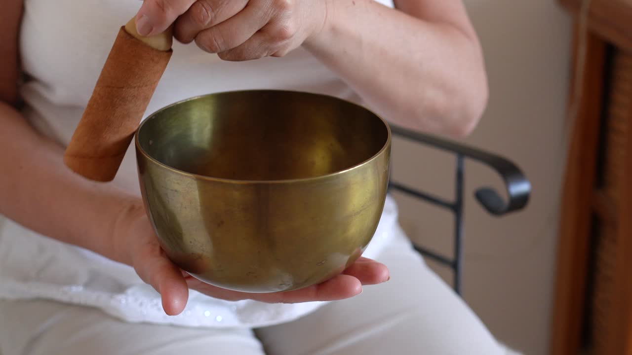 Seated Woman Rotating Wooden Mallet Around Brass Tibetan Singing Bowl While Holding It In One Hand. closeup shot