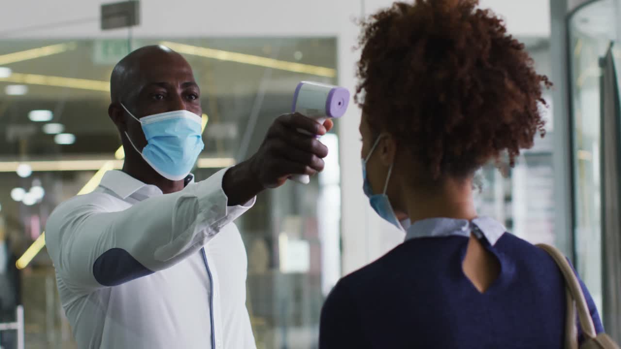 African american businessman in face mask taking temperature of female colleague arriving at office