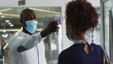 Hombre-De-Negocios-Afroamericano-Con-Mascarilla-Tomando-La-Temperatura-De-Una-Colega-Que-Llega-A-La-Oficina