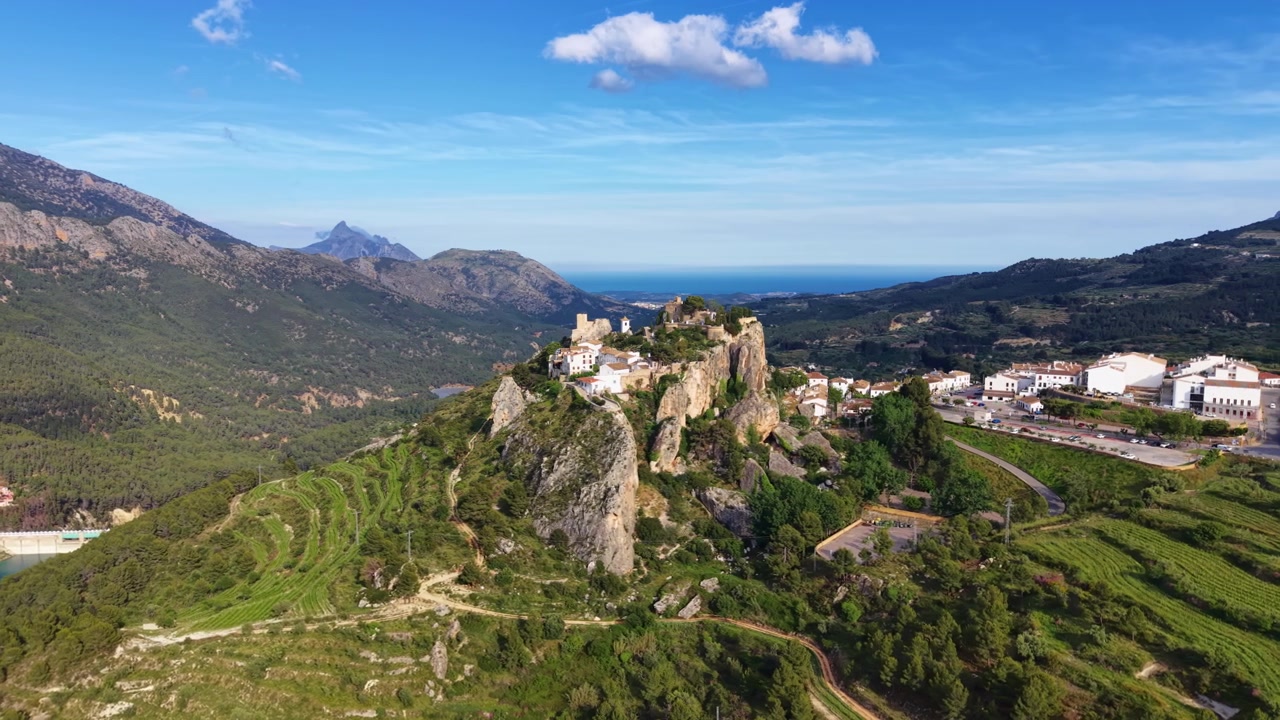 Castello di Guadalest arroccato su una collina rocciosa ad Alicante, Spagna, circondato da verdi valli montane e Mar Mediterraneo visibile sullo sfondo.