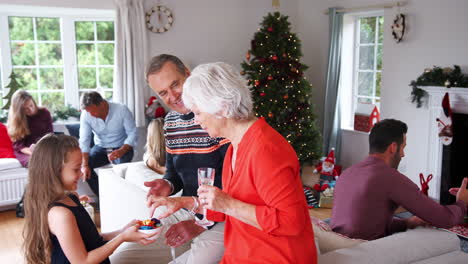 granddaughter serving sweets to grandparents as multi-generation family and friends celebrate at christmas house party