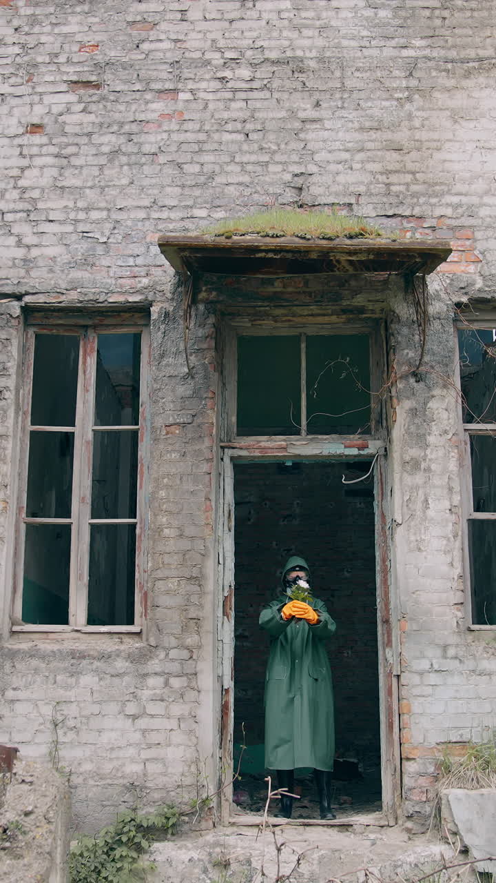 Human in infected zone. Ecologist in protective suit and gas mask coming out the abandoned building and holding flowers in hands. Vertical video