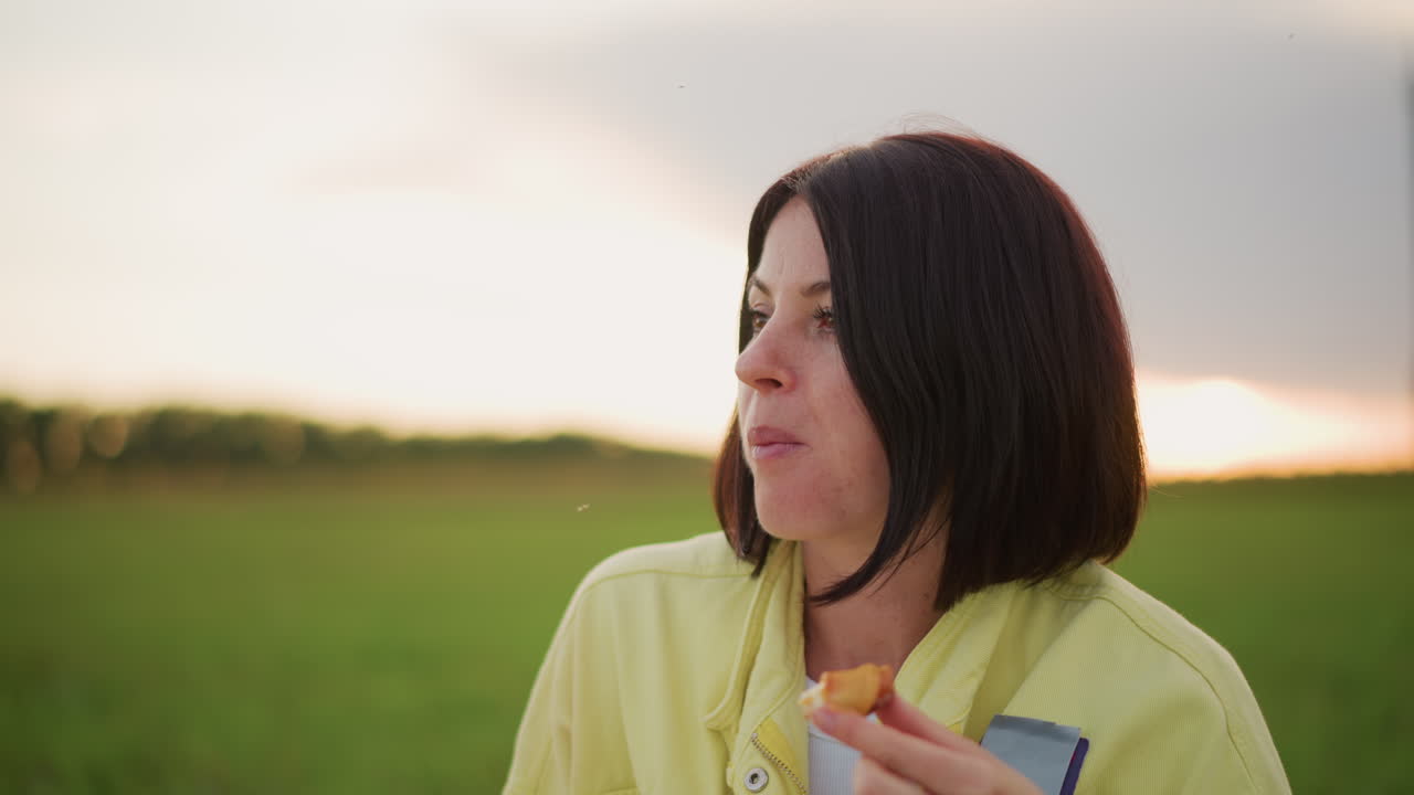 황혼에 복숭아를 먹는 백인 여성, 들판 클로즈업 인물 사진, 노란색 재킷을 입고 과일과 음료를 들고 사색에 잠긴 표정, 부드러운 황금빛, 조용한 고독, 솔직한 라이프스타일 영상
