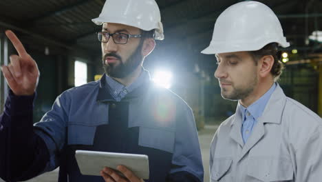 deux ouvriers portant des casques parlent en regardant la tablette dans une grande usine