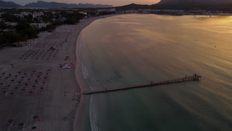 Vista-Aérea-De-La-Playa-De-Alcúdia,-Mallorca-Durante-El-Amanecer-Con-El-Muelle-Y-Las-Montañas-De-Fondo