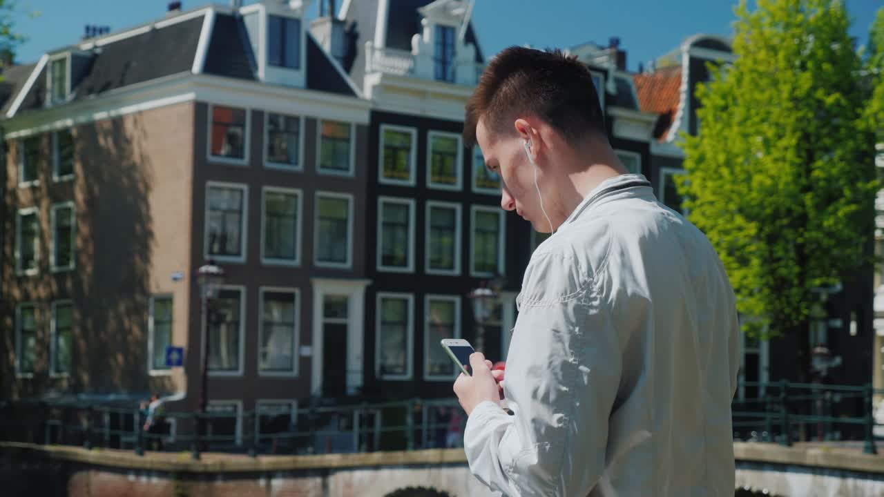 A teenager listens to music on headphones, holds a smartphone in his hand. It is near the bridge across the canal in Amsterdam. Tourism and study in the Netherlands concept