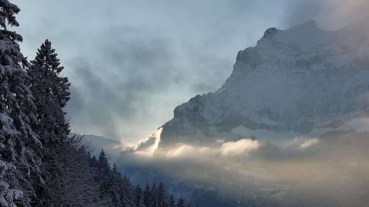 Aerial view of Rautispitz peak in the Glärnisch mountain range, Glarus, Switzerland. Sunlight pierces through clouds, casting a golden glow over the snow-covered alpine landscape.