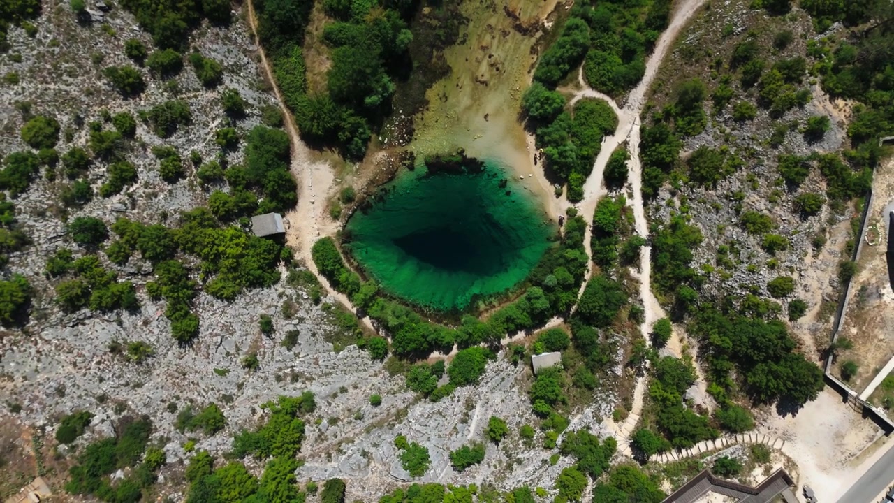 Riprese aeree con drone che rivelano una sorgente carsica turchese cristallina, circondata da un terreno calcareo accidentato, vegetazione verde e sentieri. Paesaggio naturale scenografico ideale per viaggi e turismo.
