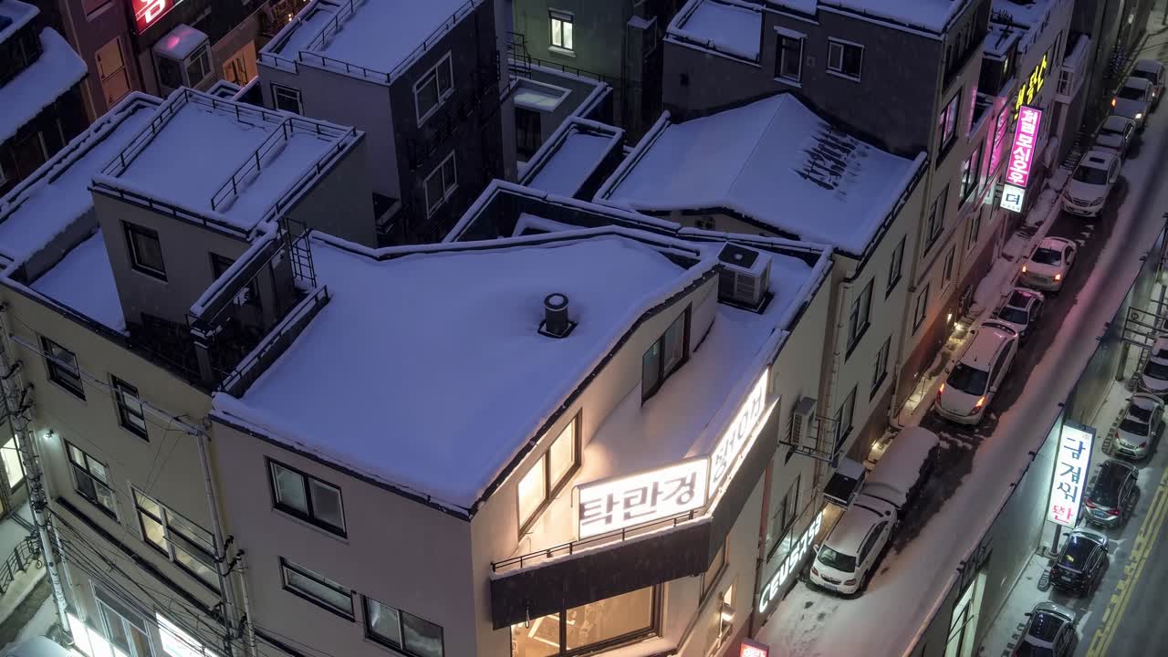 Snow is covering the rooftops of buildings in an urban area of Korea, cars are parked along the street, and neon signs add a touch of color to the dusky scene