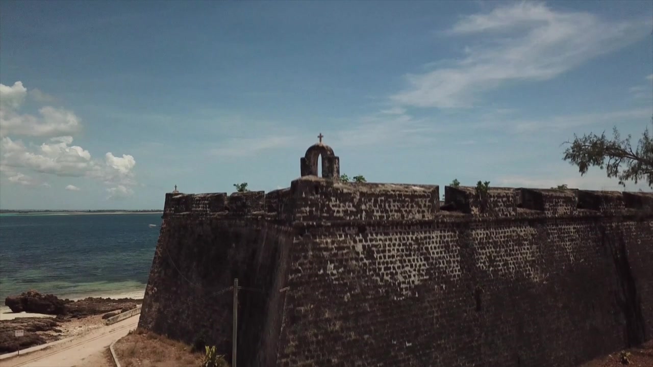 Lenta ascesa verticale di un drone che rivela gli storici bastioni del Forte di São Sebastião, sormontati da un arco in pietra con una croce, che si affacciano sull'Oceano Indiano turchese sull'Isola del Mozambico.