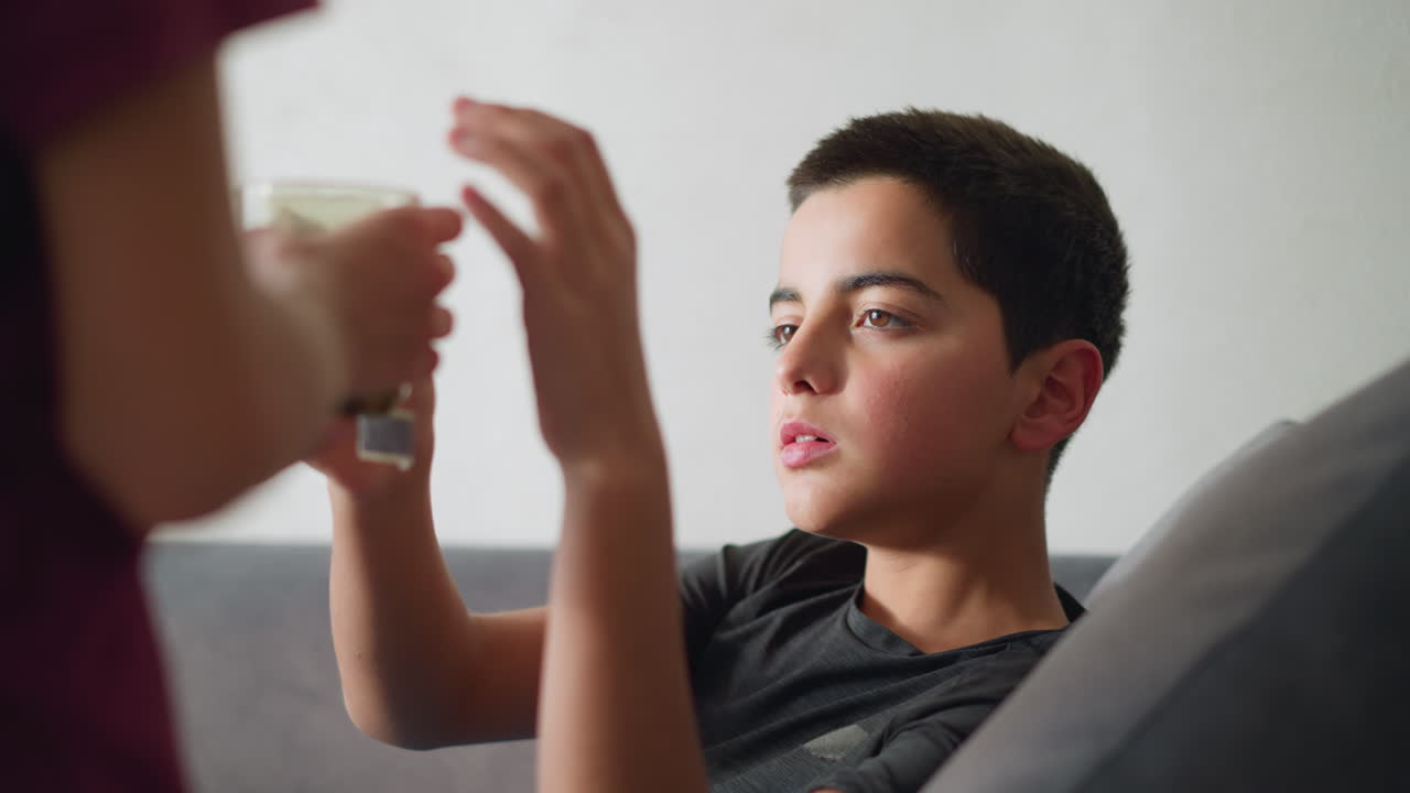 Side view of young girl handing herbal tea to her brother seated on couch, offering care and comfort during illness as he collects and drinks from the cup