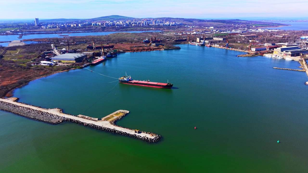 Vista panoramica di una baia portuale industriale, Bulgaria. Ampia veduta aerea con drone che mostra una nave da carico rossa circondata dall'acqua, strutture frangiflutti e una città costiera in lontananza.