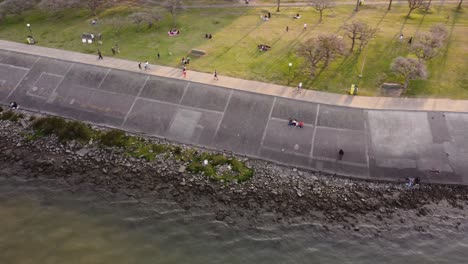 Toma-Aérea-De-Personas-Relajándose-En-La-Pradera,-Montando-En-Bicicleta-Y-Caminando-A-Lo-Largo-De-La-Orilla-Del-Río-De-La-Plata,-Buenos-Aires