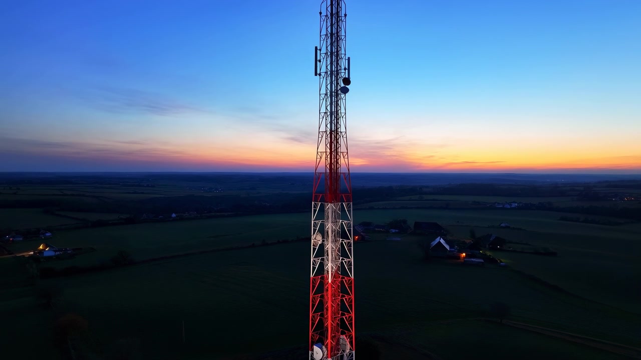 Tall red and white lattice telecommunications tower stands above rural farmland in sunset light, with mounted panel antennas, parabolic dishes, open sky, distant horizon, and warm evening glow