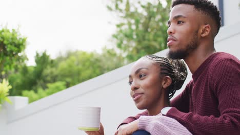 Happy-african-american-couple-embracing-and-relaxing-with-coffee-outside-house
