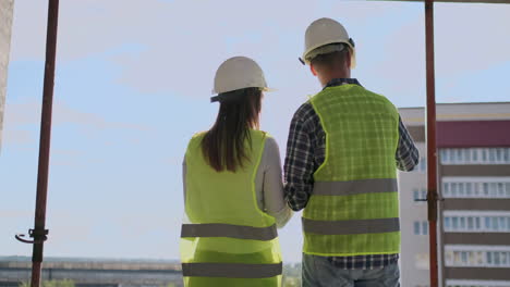 Vista-Posterior,-Joven-Capataz-E-Ingeniero-Revisando-El-Plano-Y-Hablando-Sobre-El-Proyecto-De-Construcción-Con-Compromiso-Con-El-éxito-En-El-Sitio-De-Construcción