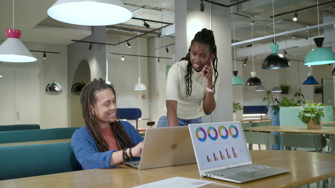 African American coworkers during phone call reviewing charts at table, pointing at laptop, papers