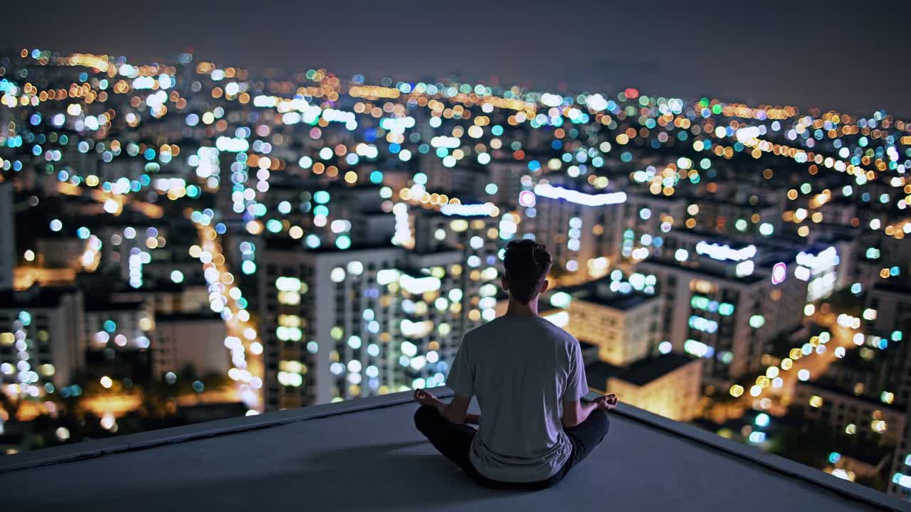 Individual in a meditative pose on a rooftop at night, overlooking a vibrant cityscape filled with shimmering lights, embodying tranquility and urban serenity