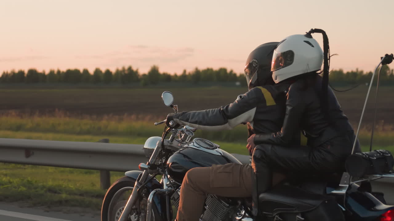Man and woman wearing helmets ride motorcycle along countryside road past fields during sunset, passenger holding rider tightly as they drive past kilometer signpost