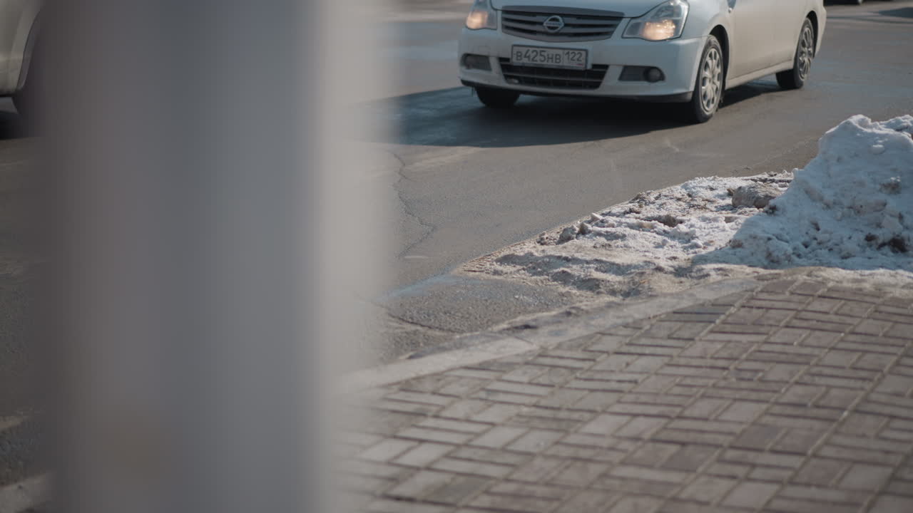 busy city view shows cars moving along winter road past snow pile on corner, cracked asphalt meets brick pavement near curb, sun glints on wet surface, foreground pole adds subtle frame