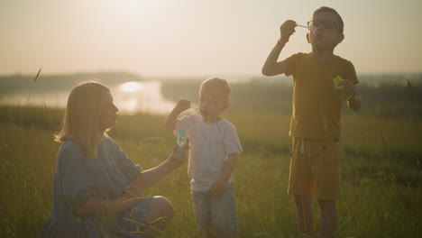 joyful woman in a blue dress kneels in a grassy field at sunset, holding a bubble bottle as her younger son dips a wand and blows bubbles. her older son, dressed in a yellow outfit, also blows bubbles