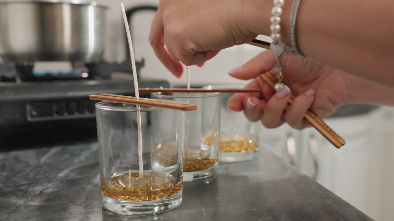 Close up of person using wooden chopsticks to secure candle wick firmly at center of glass cups filled with melted wax beads, with background view of pot on gas burner in modern kitchen setting