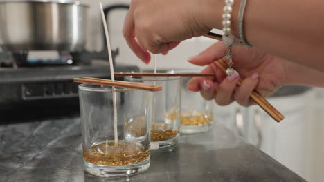 close up of person using wooden chopsticks to secure candle wick firmly at center of glass cups filled with melted wax beads, with background view of pot on gas burner in modern kitchen setting