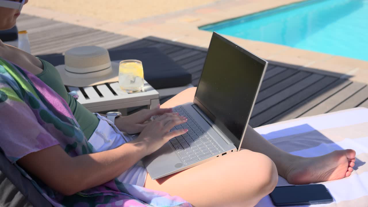 Donna che si sistema su una sedia a sdraio, lavora e digita sul laptop a bordo piscina, allungando la mano per una bibita e sorridendo.