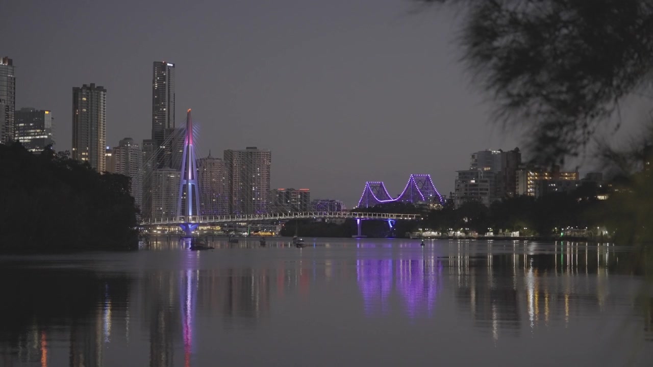 Una vista notturna sul fiume Brisbane mostra il Ponte Kangaroo Point illuminato di viola sotto uno skyline cittadino illuminato