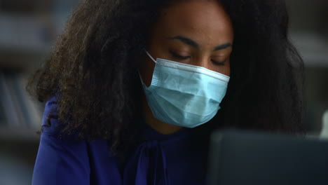 African-american-businesswoman-wearing-mask-searching-laptop-computer-in-office.