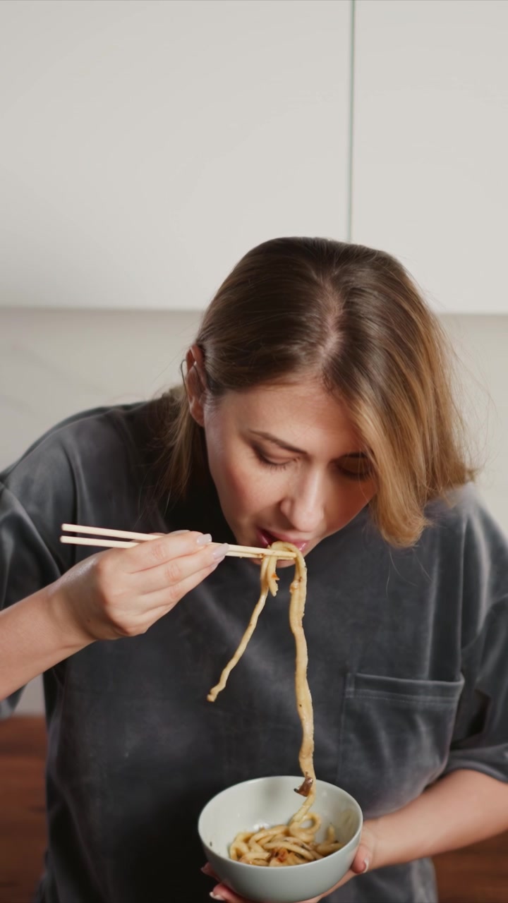 Primo piano di una donna che usa le bacchette per mangiare noodles da una ciotola. La scena è ambientata al chiuso con illuminazione soffusa, che si concentra sull'azione e sull'interazione tra mani, utensili, la consistenza del cibo, i dettagli e un'atmosfera calma.