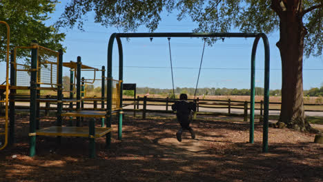 a young child with her back turned swinging on a swing on a playground with a road in the background and surrounded by trees