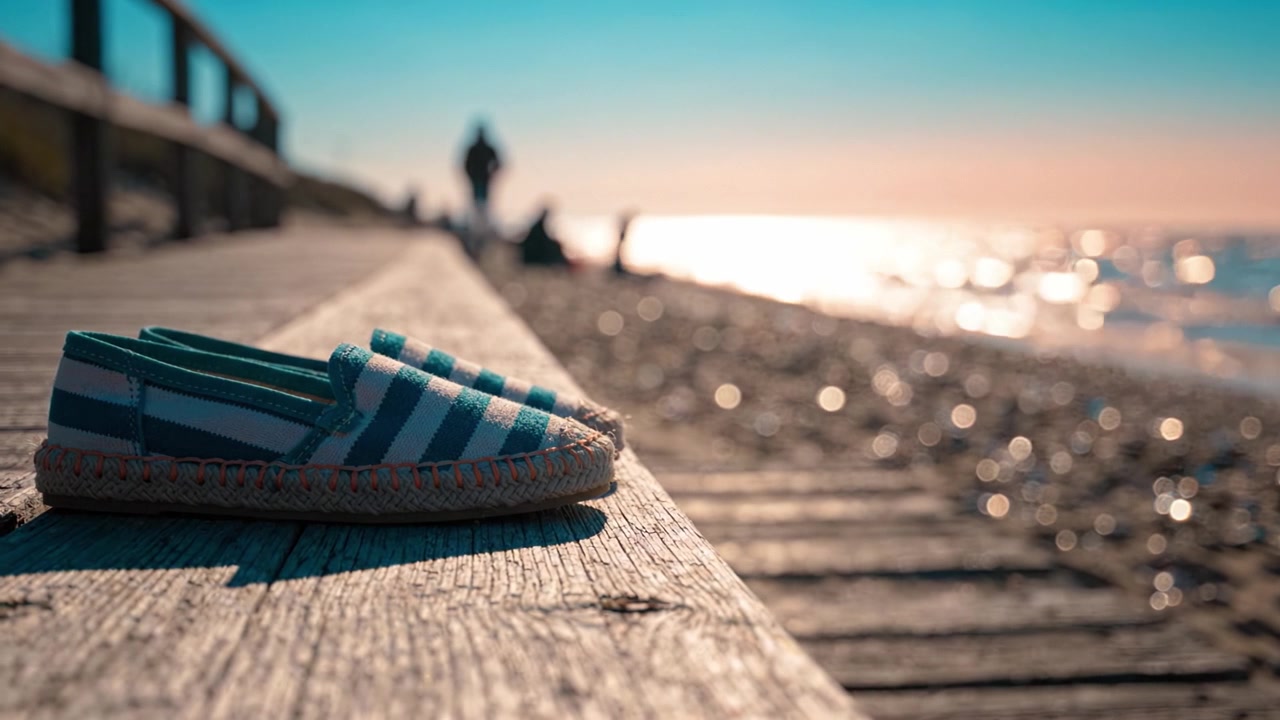 Une paire d'espadrilles rayées reposant sur une promenade en bois patinée au bord de l'eau, les vagues océaniques scintillantes et la lumière du soleil en arrière-plan créent une atmosphère sereine.