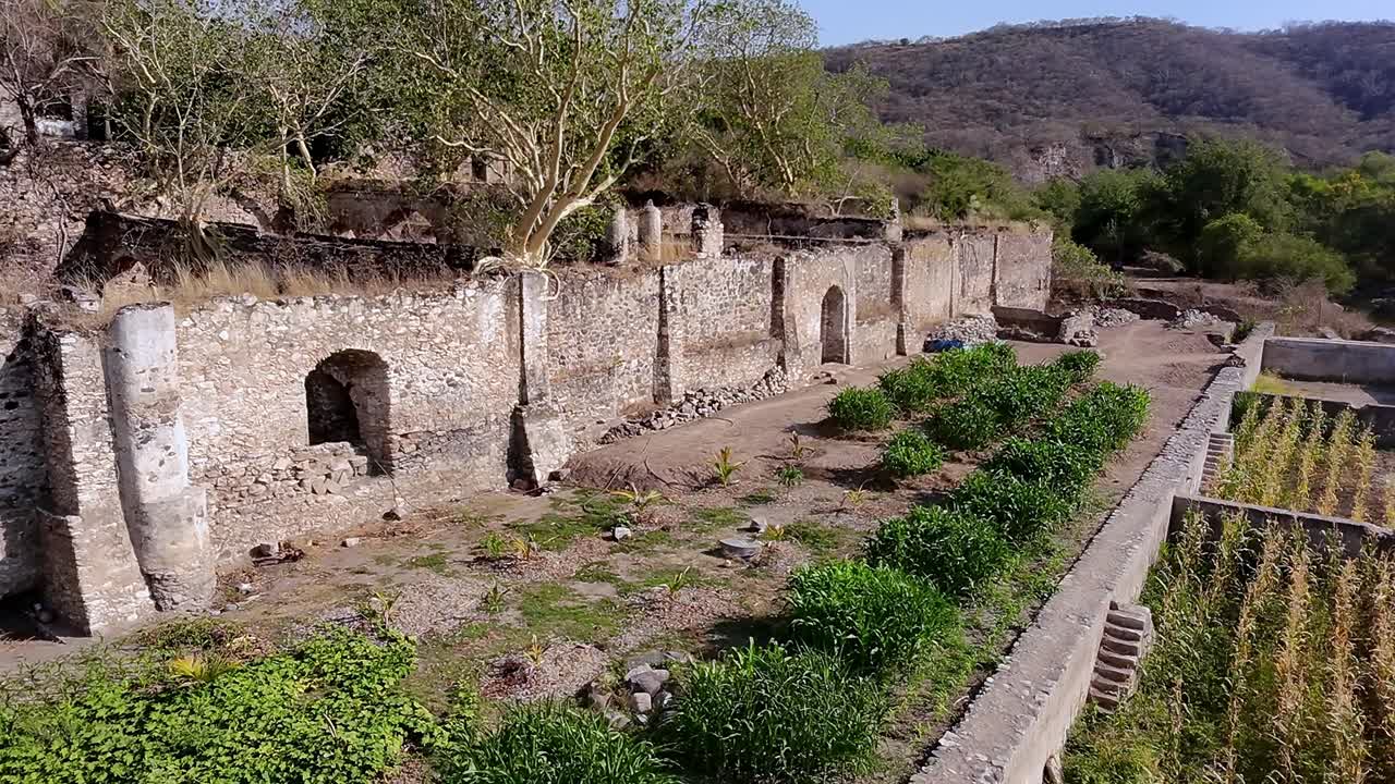 Backward drone flight reveals Ex Hacienda San Jacinto Ixtoluca, La Mezquitera, Morelos. Stone ruins of an abandoned estate with arched structures, trees, and cultivated greenery