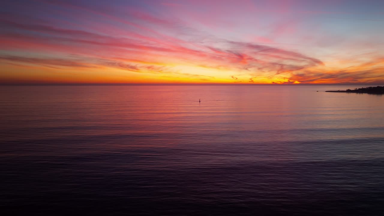 A beautiful and colorful ocean sunset as seen from Santa Cruz California looking over the pacific ocean towards a lone sailboat in a serene panning aerial drone shot