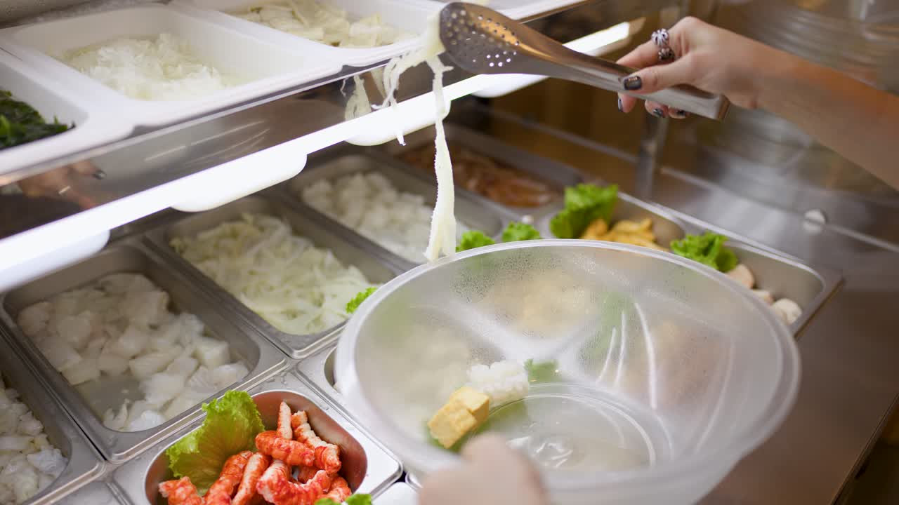 Person uses tongs to pick seafood and vegetables from buffet trays under bright lighting