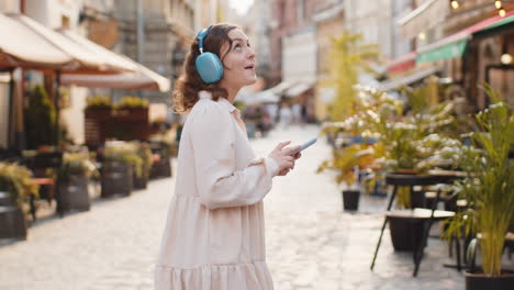 happy young woman in wireless headphones choosing, listening music in smartphone dancing outdoors