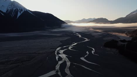 Luftaufnahme-Des-Geflochtenen-Bealey-River-Im-Schatten-Im-Arthurs-Pass-Bei-Sonnenaufgang-In-Einem-Von-Bergen-Umgebenen-Tal,-In-Richtung-Der-Berge-Und-Der-Sonne-Fliegend