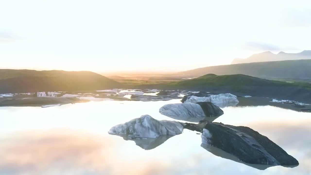Aerial drone flying backwards revealing gorgeous reflective water in the middle of icy white glaciers in Iceland or Greenland during a gorgeous sunny day. Sky and icebergs are reflecting on cold water