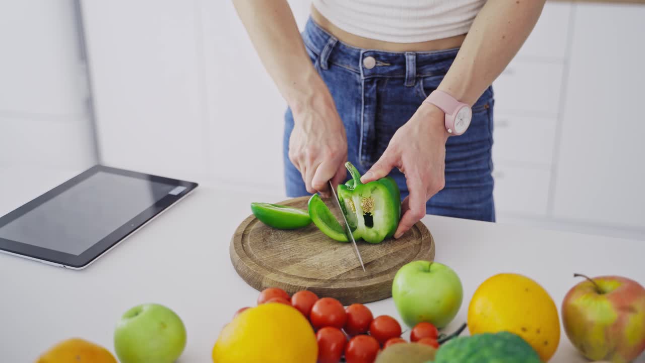 Young female is cutting green pepper on a wooden board. Close-up woman hands preparing healthy food on the kitchen on fresh ingredients background.